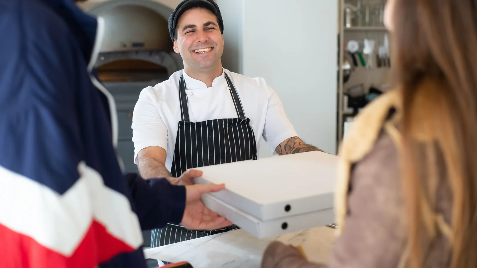 Chef entregando cajas de pizza a clientes en una pizzería acogedora con ambiente cálido | ZonaFranquicia