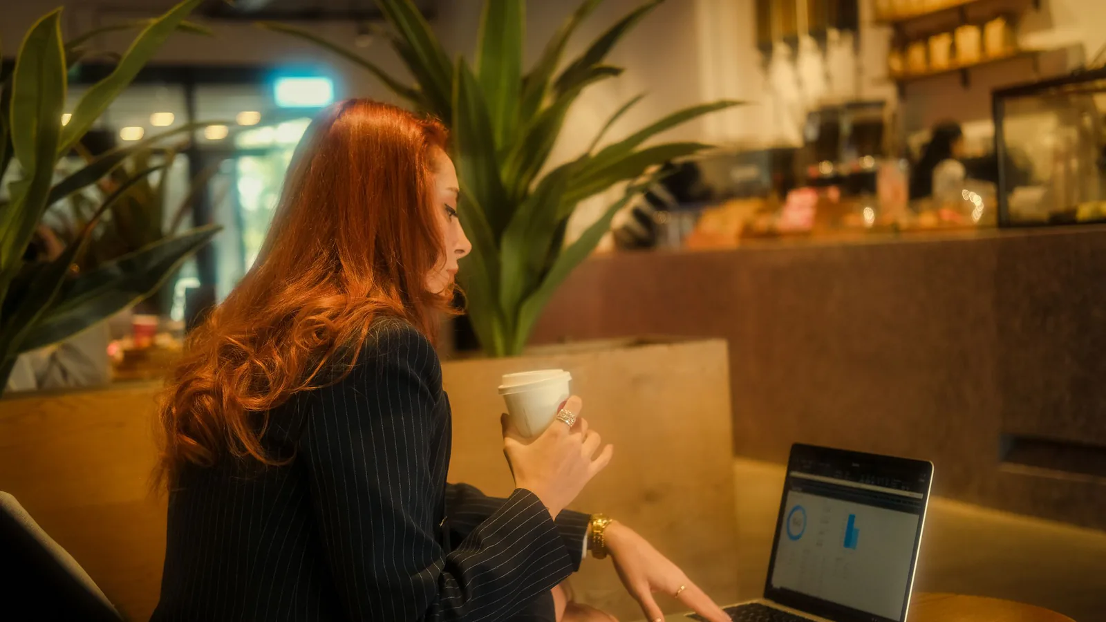 Franquicia a distancia: gestiona tu negocio sin estar allí | ZonaFranquicia Empresaria pelirroja trabajando en laptop en cafetería acogedora, sosteniendo un café | ZonaFranquicia