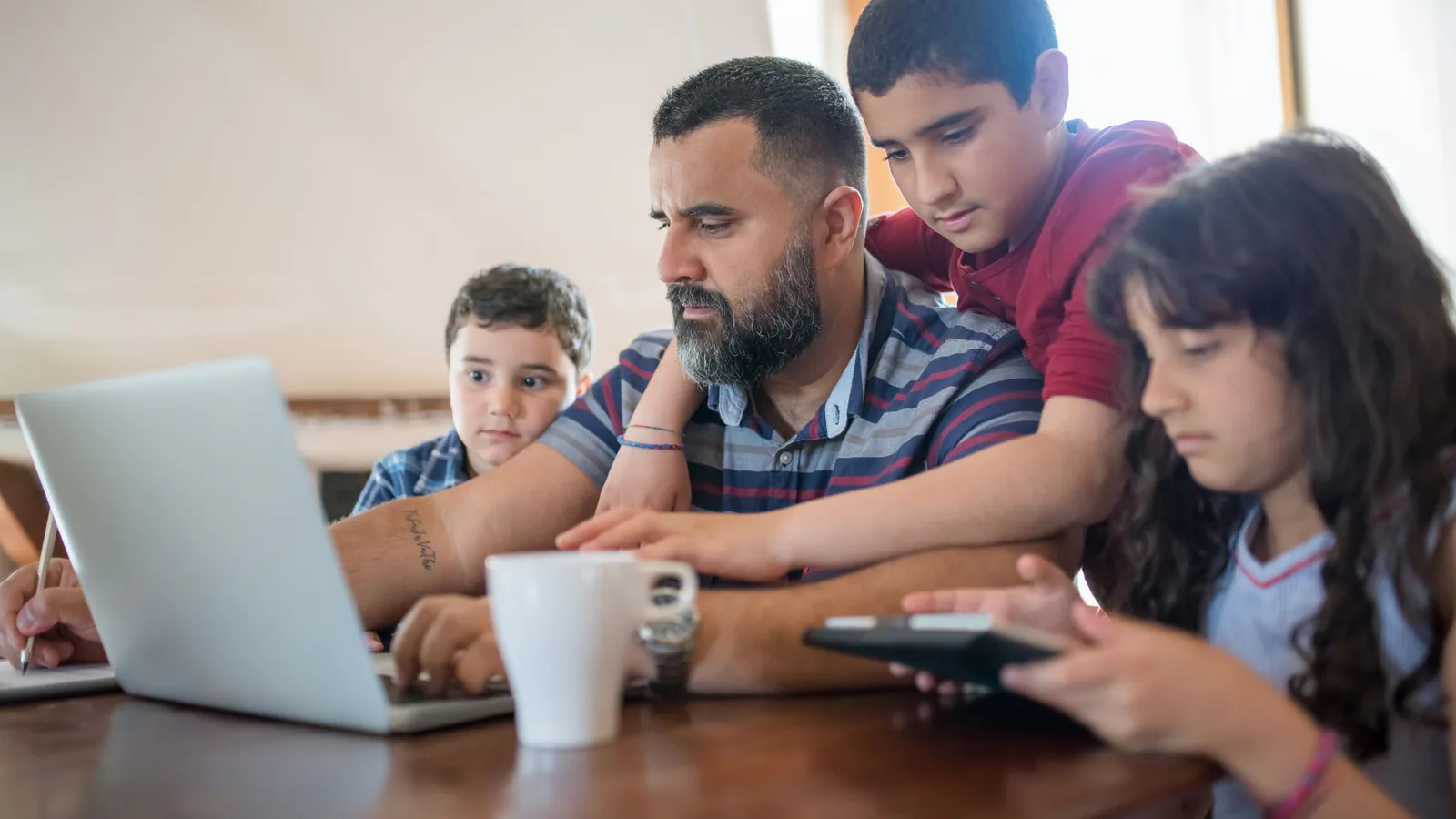 Franquicia familiar: cómo involucrar pareja e hijos sin romper | ZonaFranquicia Padre trabajando en laptop mientras colabora con sus hijos alrededor de la mesa en casa | ZonaFranquicia