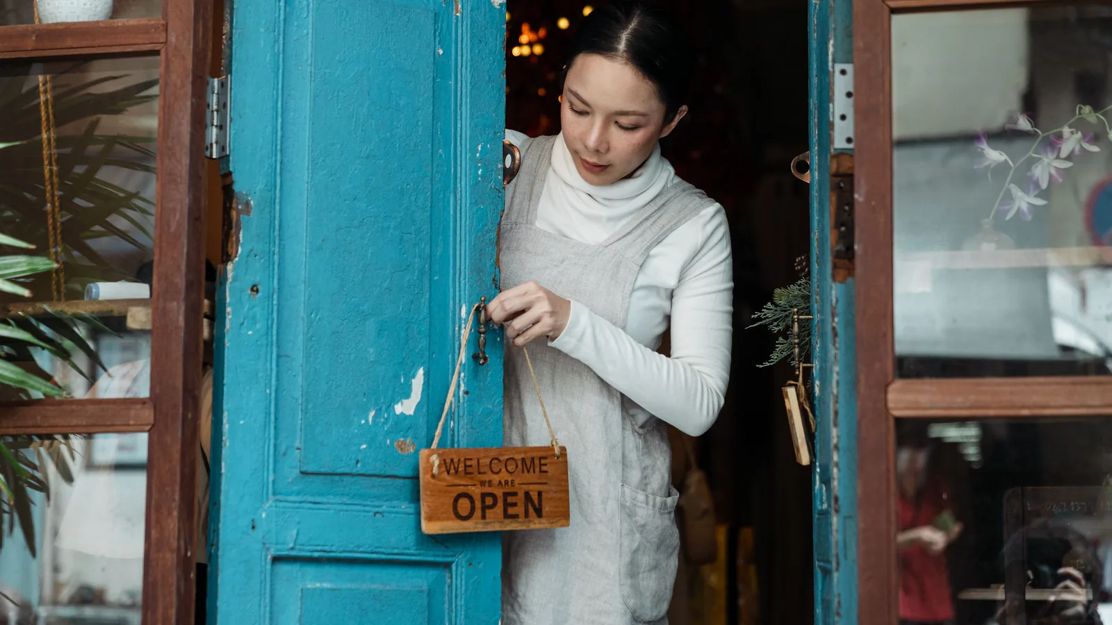 La franquicia española resiste: factura un 3% más pese a tener menos marcas y locales | ZonaFranquicia Una mujer ajusta el cartel de abierto en la puerta de madera azul de una cafetería. | ZonaFranquicia