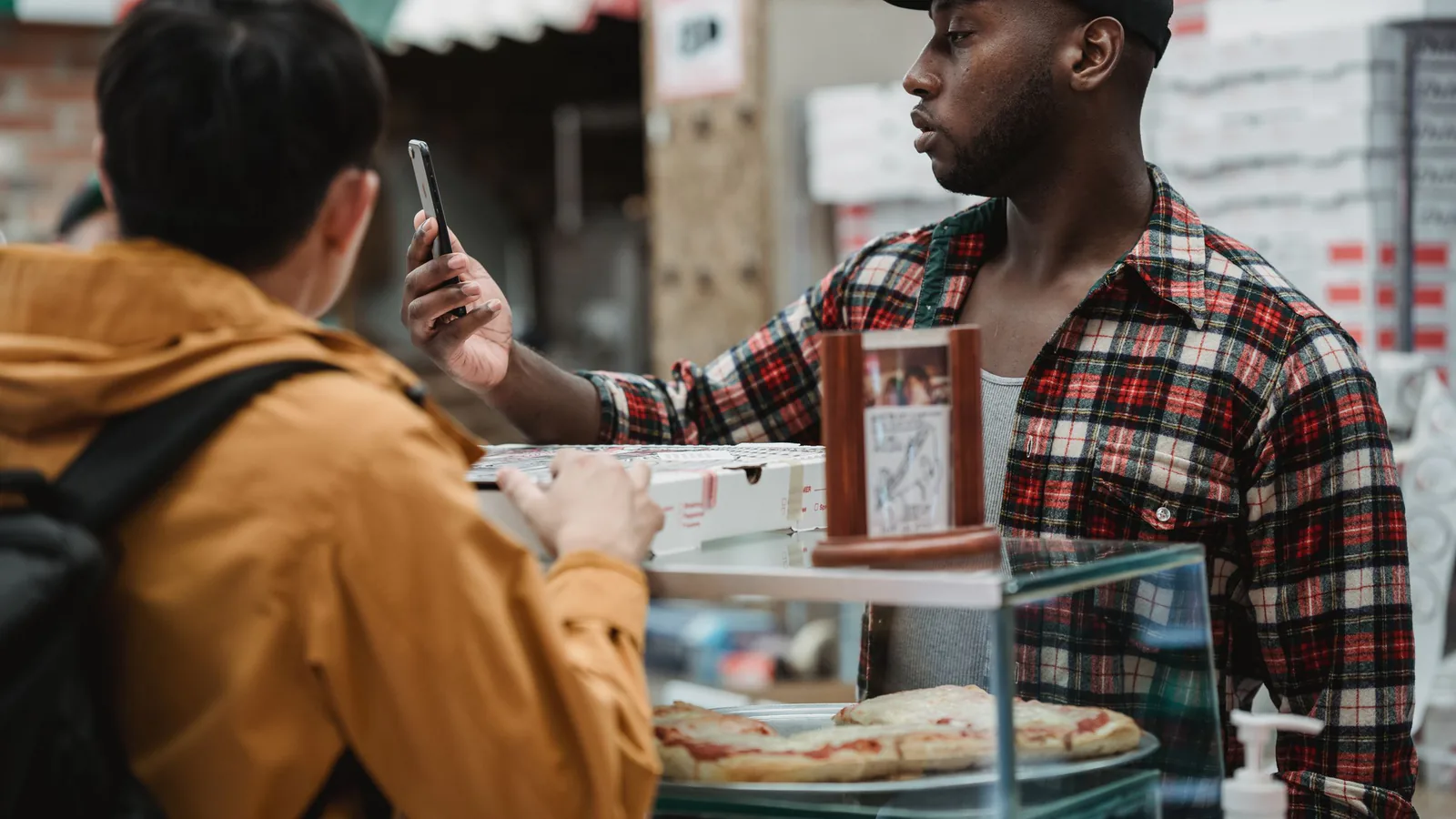 Pizzacadabra abre su pizzería automática 24 horas en Madrid y suma otra franquicia al modelo de las… | ZonaFranquicia Dos personas interactuando en el mostrador de una pizzería moderna, una de ellas con smartphone | ZonaFranquicia